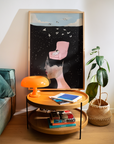 Framed art print in a cool interior setting with an orange lamp, house plant, modern furnishings.