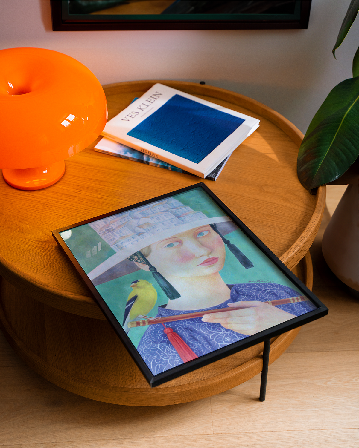 Table with a framed artwork of a woman holding a bird, an orange lamp, and books.