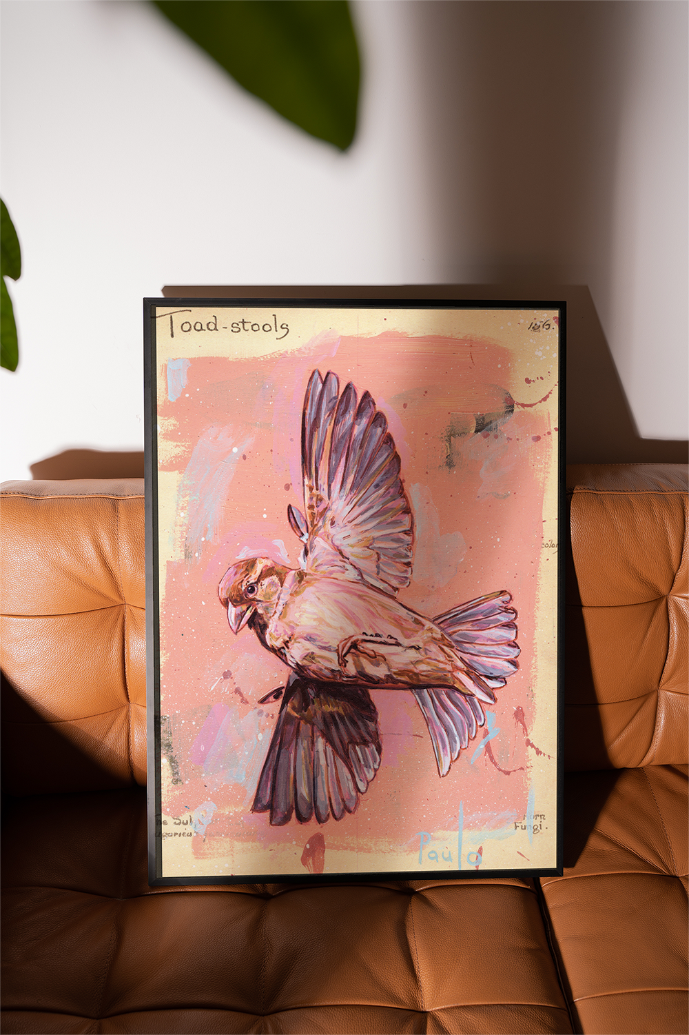 Framed artwork of a bird on a pink background, leaning against a brown leather couch.