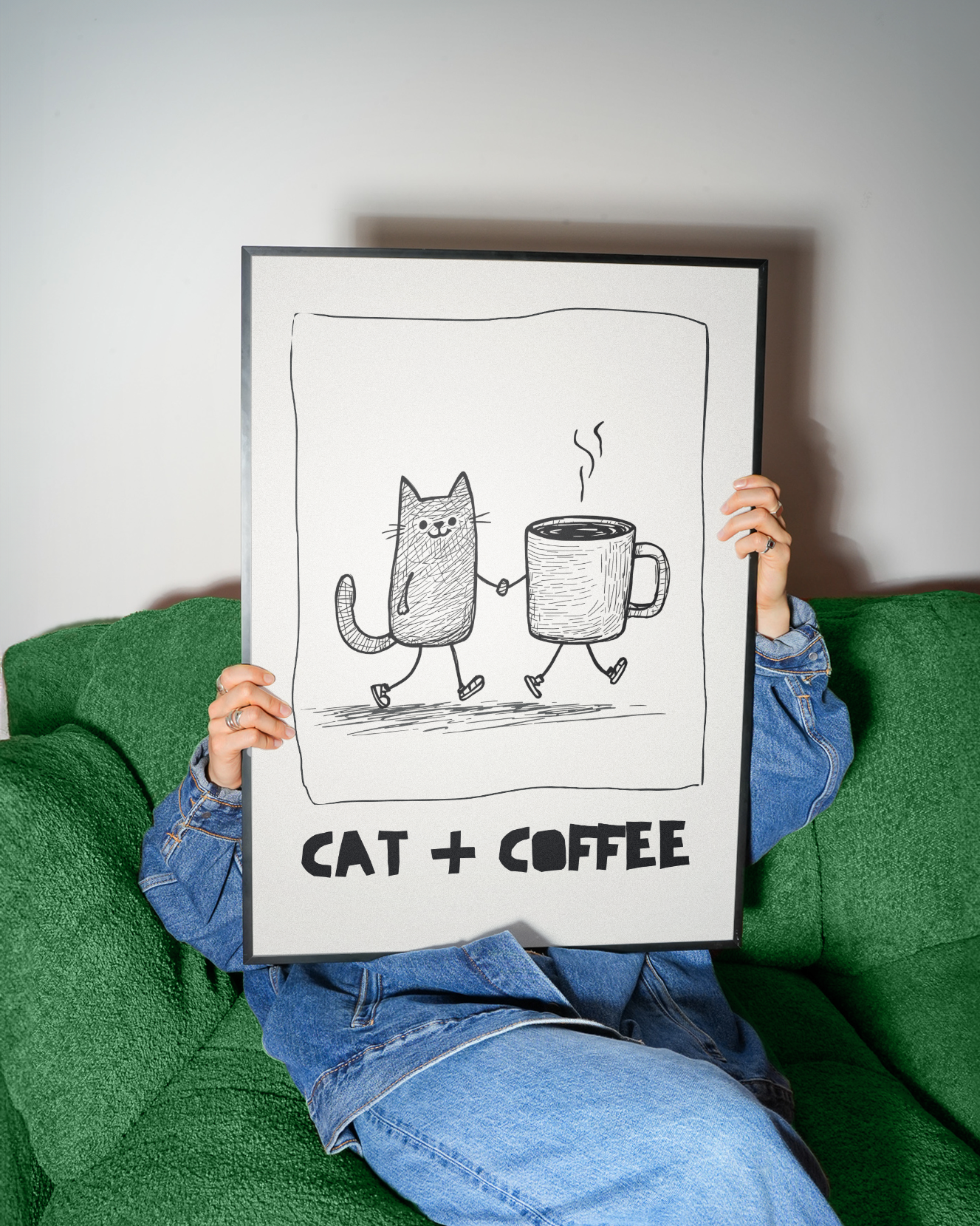 Person holding a framed illustration of a cat and coffee cup with 'CAT + COFFEE' text on a green couch.
