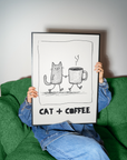 Person holding a framed illustration of a cat and coffee cup with 'CAT + COFFEE' text on a green couch.