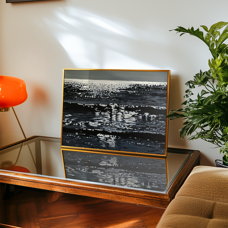 Framed ocean artwork of a sparkly sea on a table with a lamp and plant in the background