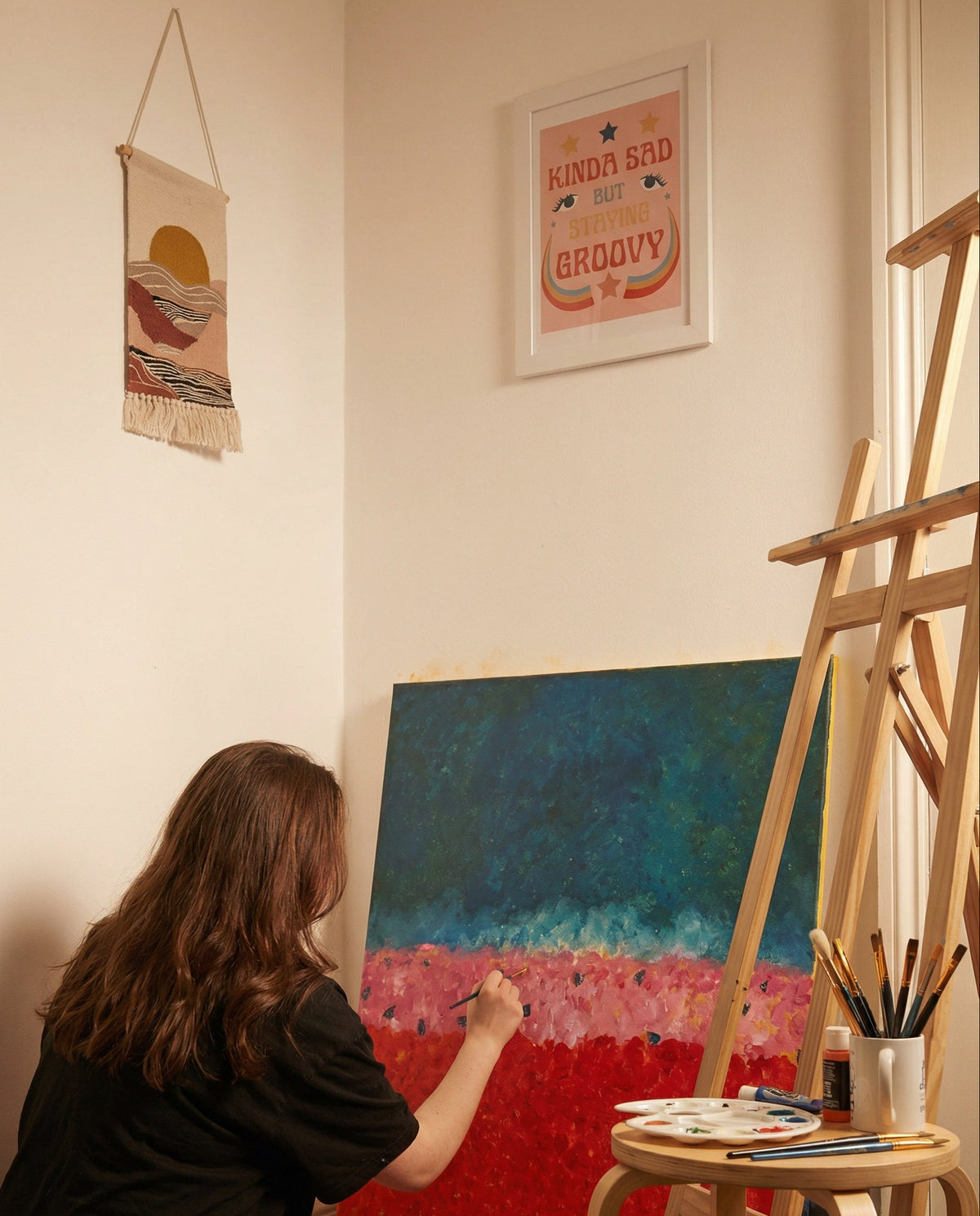 Person painting a colorful abstract scene on an easel in a room with wall art.