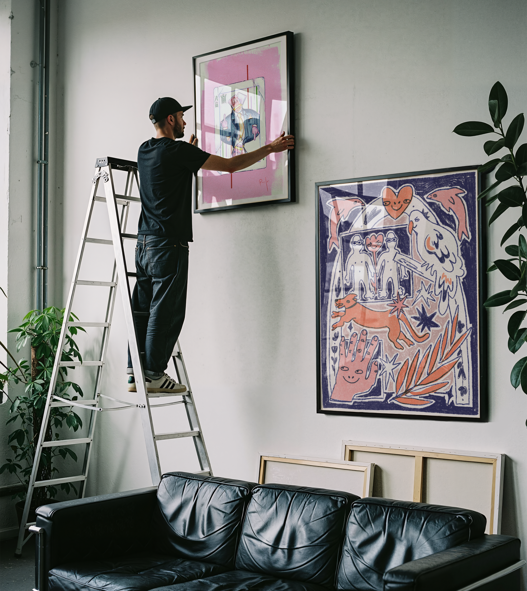 Person on a ladder hanging artwork in a living room with a black leather sofa and plants.